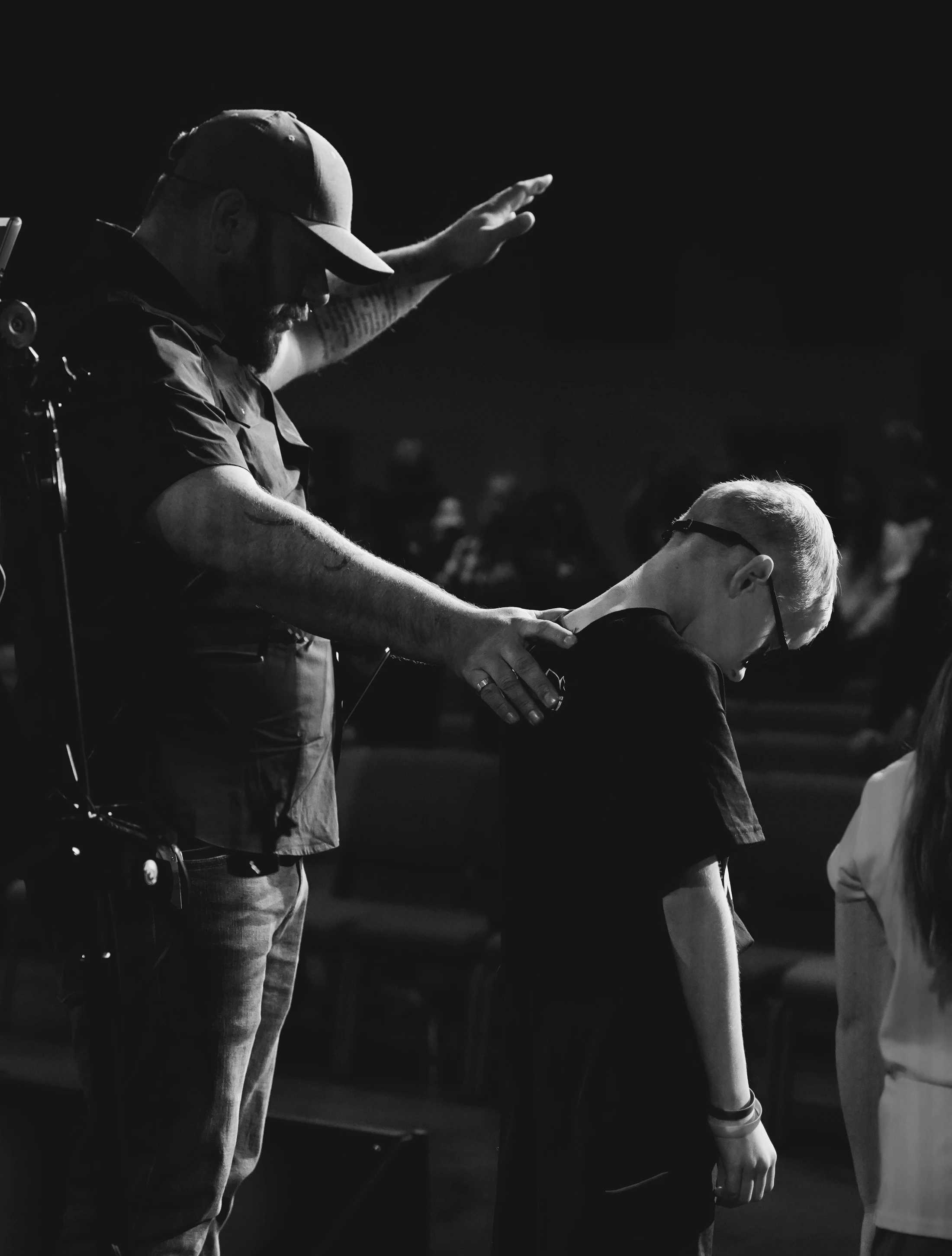 A black and white photo of a man in a baseball cap placing a hand on a young boy's shoulder and raising his other hand in a gesture of prayer or blessing.